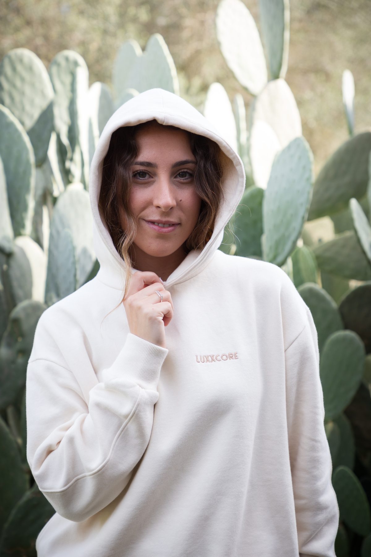 Mujer con sudadera algodón orgánico LUXXCORE ecru y capucha puesta, posando frente a cactus con mirada segura.
