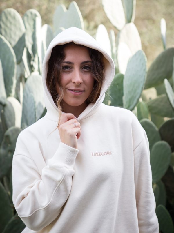 Mujer con sudadera algodón orgánico LUXXCORE ecru y capucha puesta, posando frente a cactus con mirada segura.