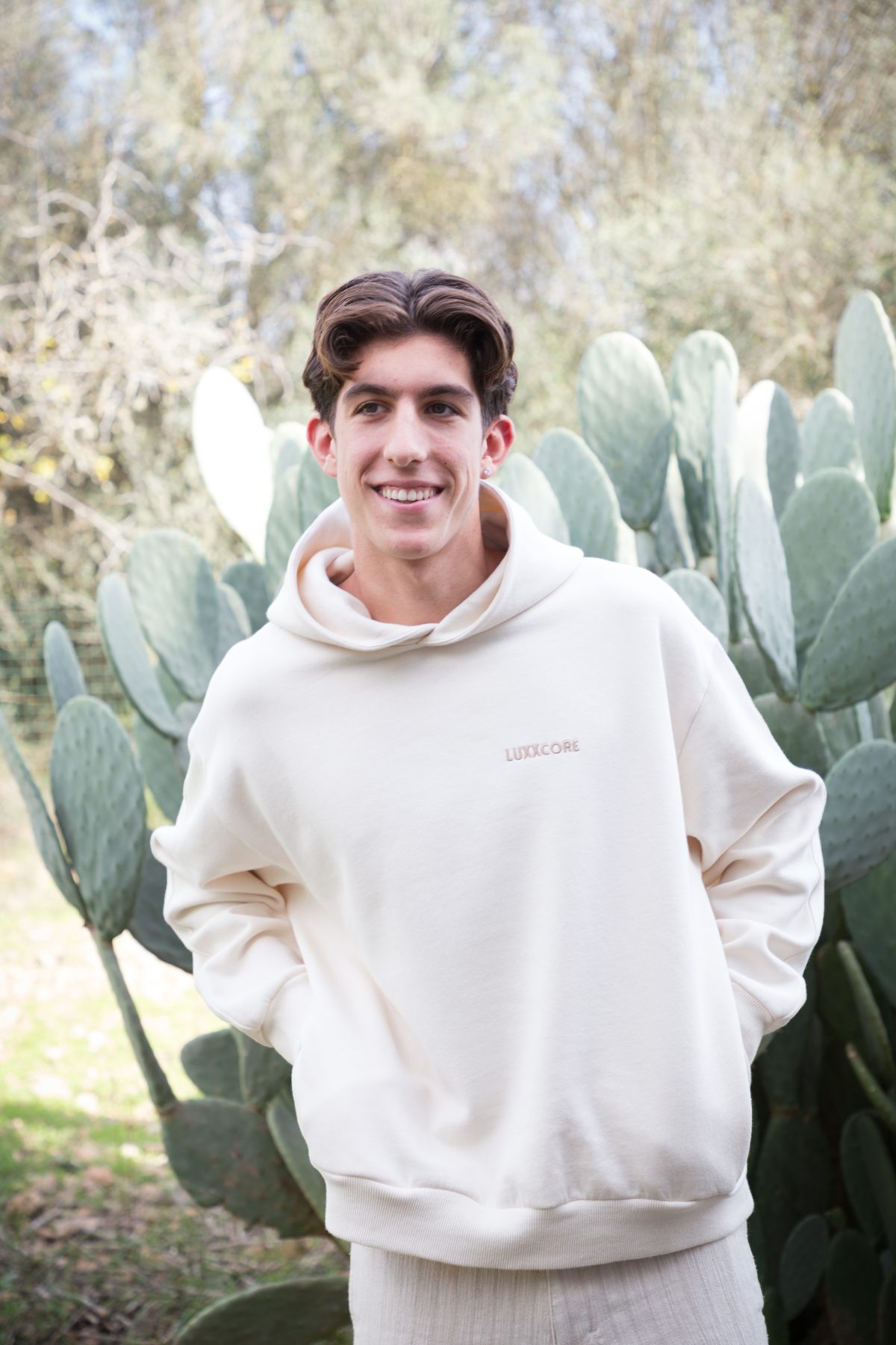 Joven con sudadera ecru algodón orgánico LUXXCORE sonriendo frente a un cactus en un entorno exterior.