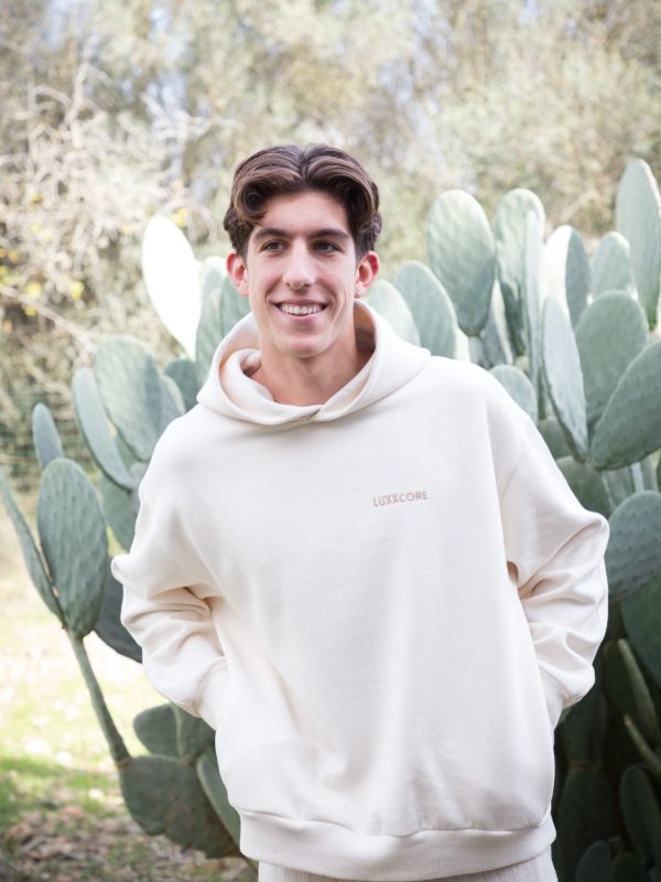 Joven con sudadera ecru algodón orgánico LUXXCORE sonriendo frente a un cactus en un entorno exterior.