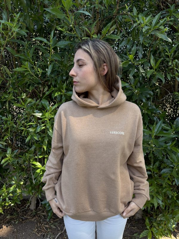 Mujer con sudadera algodón orgánico marrón LUXXCORE y capucha, de perfil, frente a un fondo frondoso de hojas verdes.