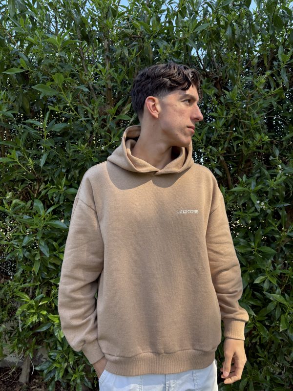 Joven con sudadera algodón orgánico marrón LUXXCORE mirando hacia un lado, frente a una vegetación frondosa.