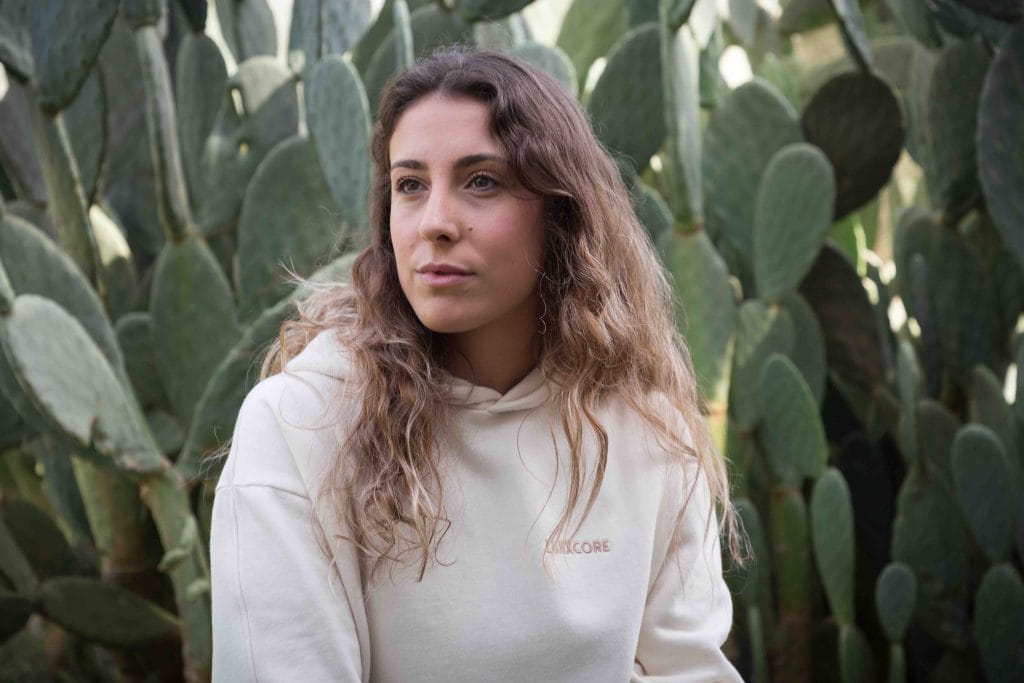 Mujer con sudadera LUXXCORE frente a un fondo de cactus, con expresión serena.