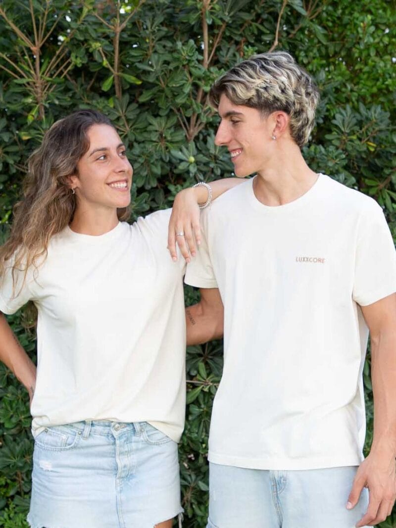 Hermana y hermano con camisetas LUXXCORE, mirándose con complicidad frente a un fondo verde natural.