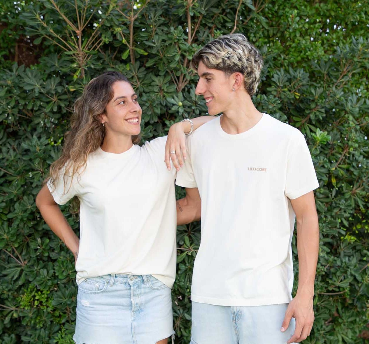 Hermana y hermano con camisetas LUXXCORE, mirándose con complicidad frente a un fondo verde natural.